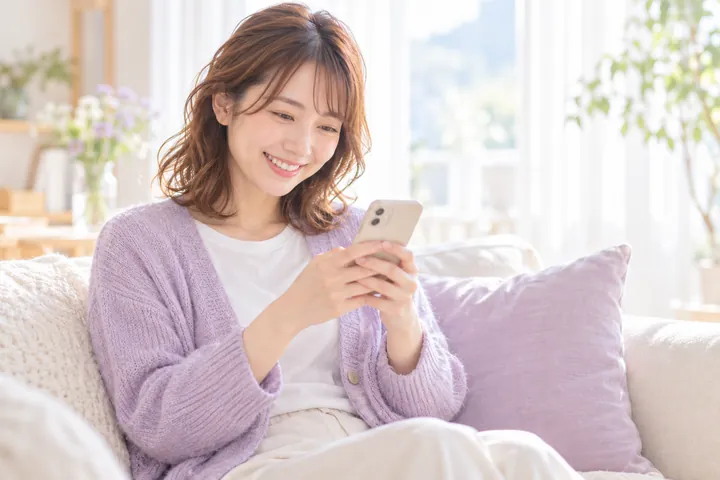 ソファでスマートフォンのメッセージを読み、ほっとした顔でほほえむ女性(AIでつくったイメージ)